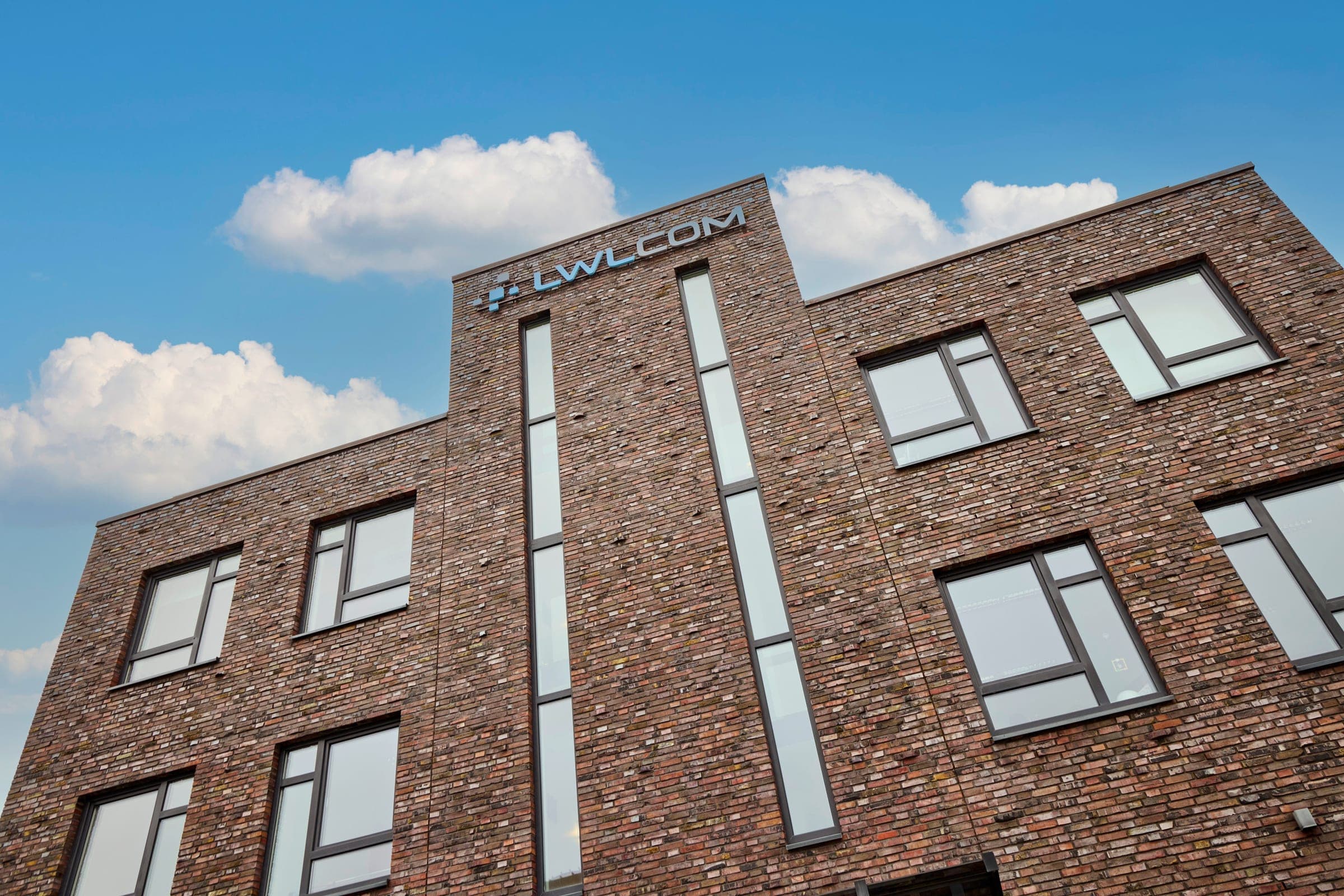 Bürogebäude der LWLcom GmbH aus schräger Untersicht, braun-rote Backsteinfassade mit mehreren rechteckigen Fenstern und LWLcom-Logo am oberen Rand.