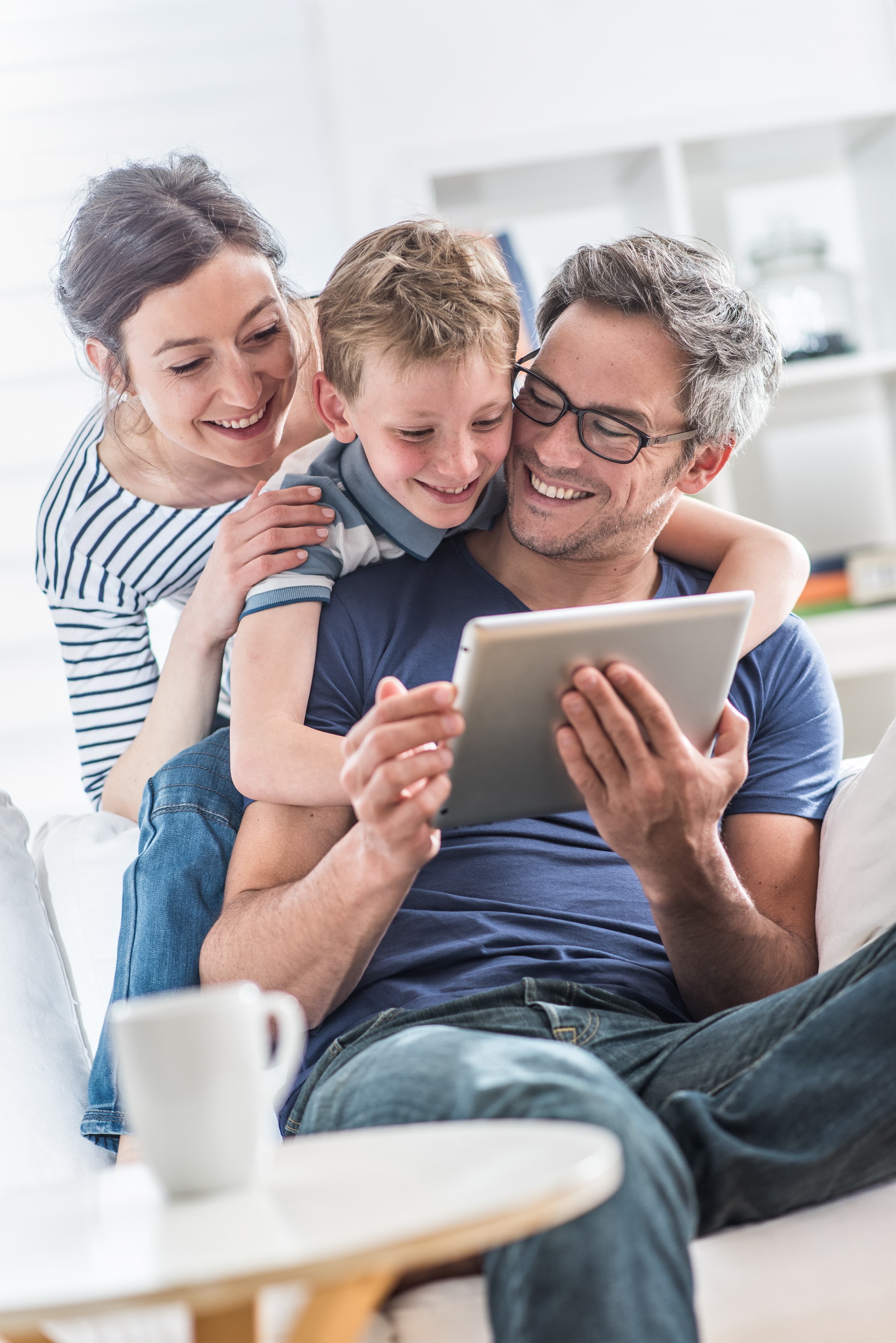 Ein Mann sitzt mit einem Tablet auf einem Stuhl. Sein Sohn und seine Frau schauen ihm über die Schulter. Alle sehen glücklich aus.