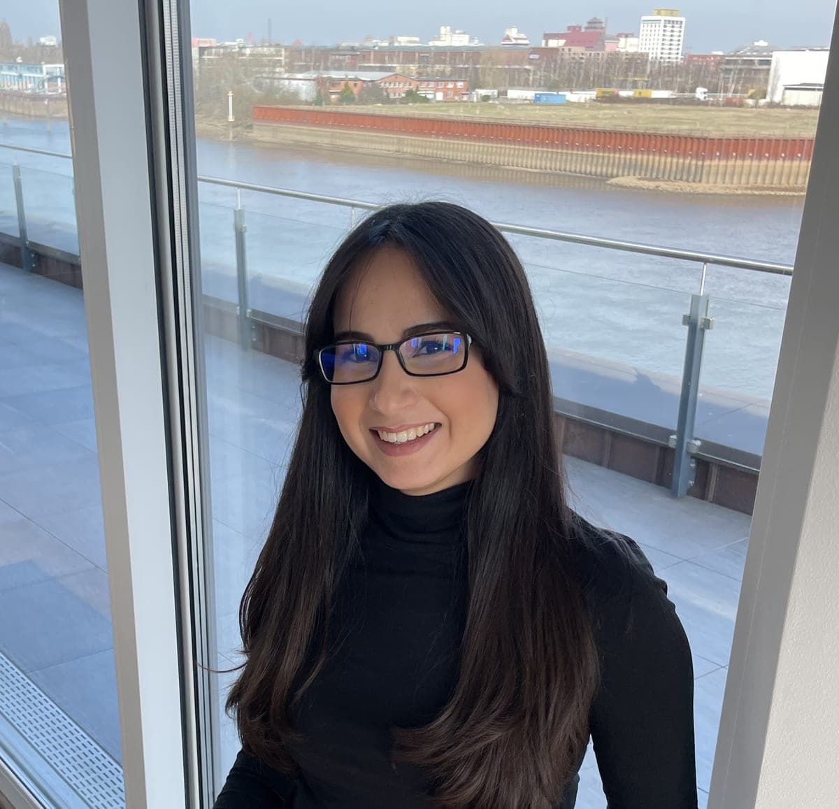 HR-Mitarbeiterin Hatice Erkekoğulları. Sie hat lange, dunkle Haare und einen schwarzen Rollkragenpullover an. Sie steht vor einem großen Fenster mit Blick auf den Fluss Weser und der Stadt Bremen im Hintergrund.