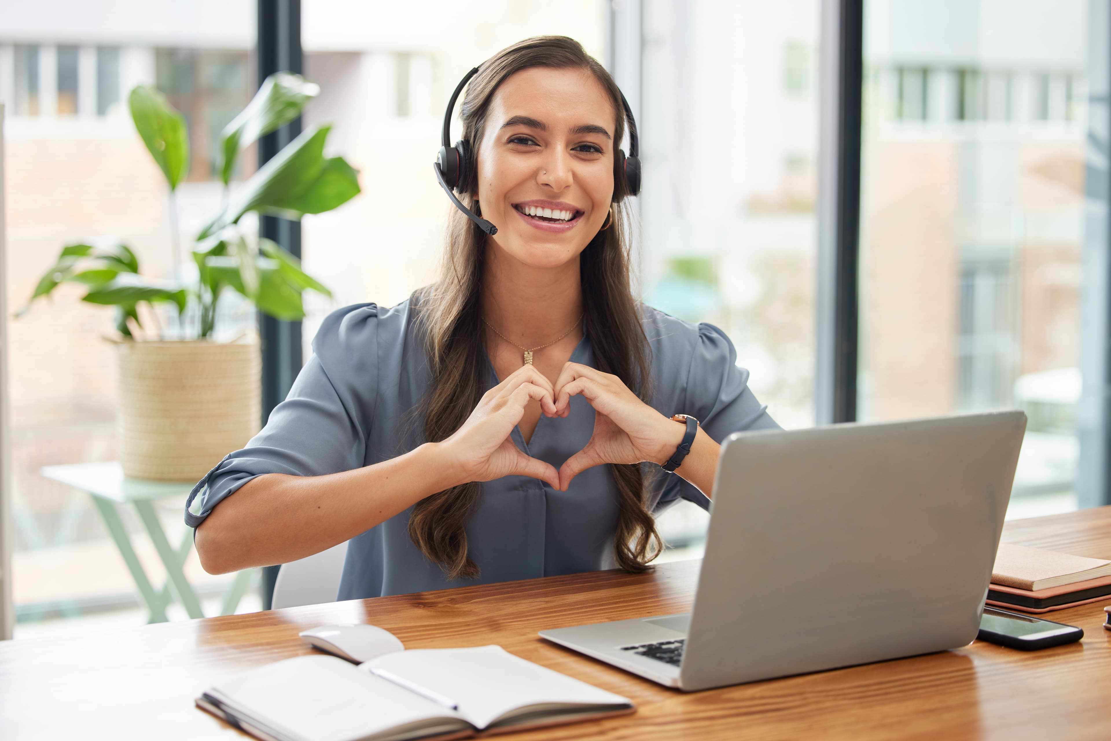 Eine Frau sitzt mit einem Laptop und Headset an einem Tisch und formt mit den Händen ein Herz.