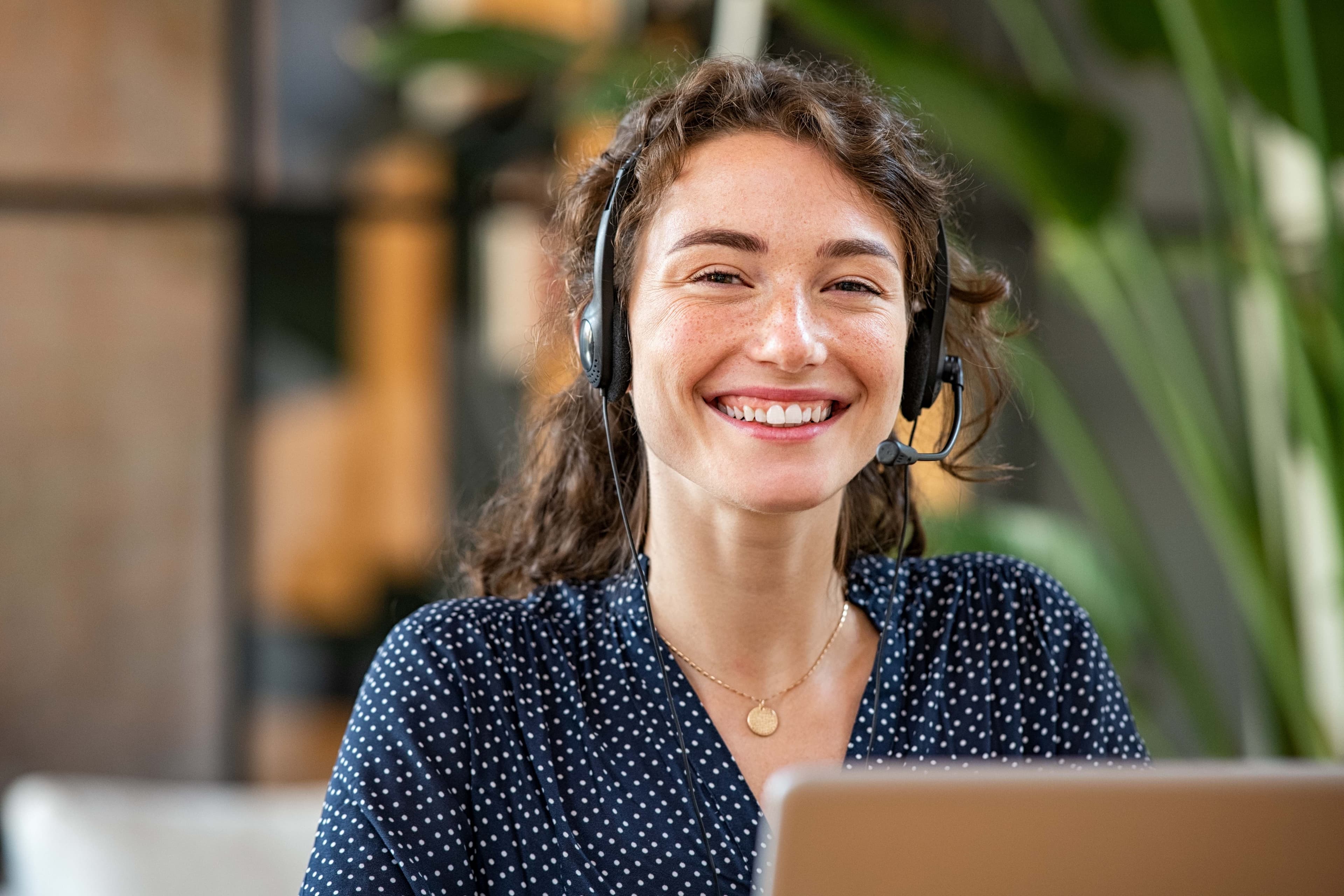 Eine Frau mit Headset sitzt vor einem Laptop und lächelt in die Kamera.