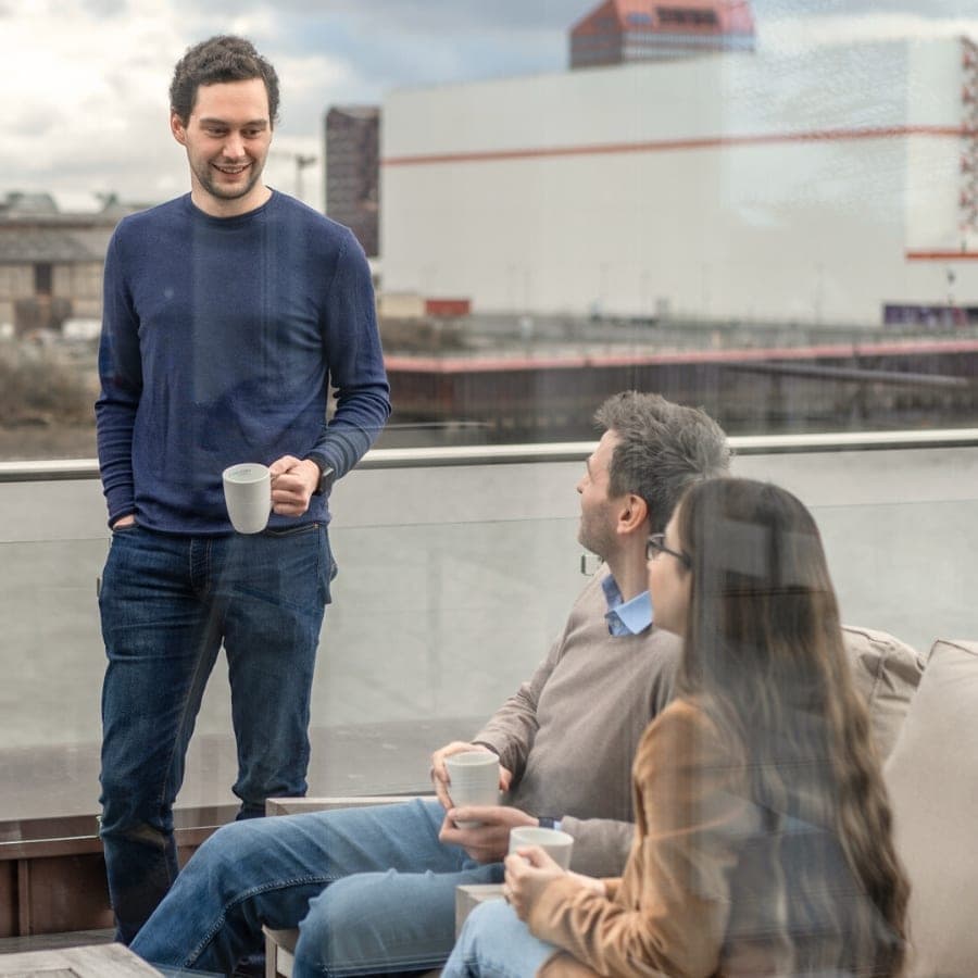 Drei Mitarbeitende mit Kaffeetassen auf der Terrasse von LWLcom, im Hintergrund die Weser