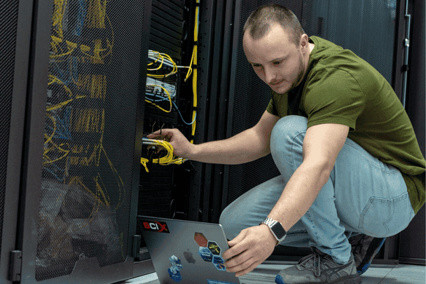 Mann mit Laptop hockt vor einem Server-RACK im Rechenzentrum und schaut konzentriert auf den Monitor.