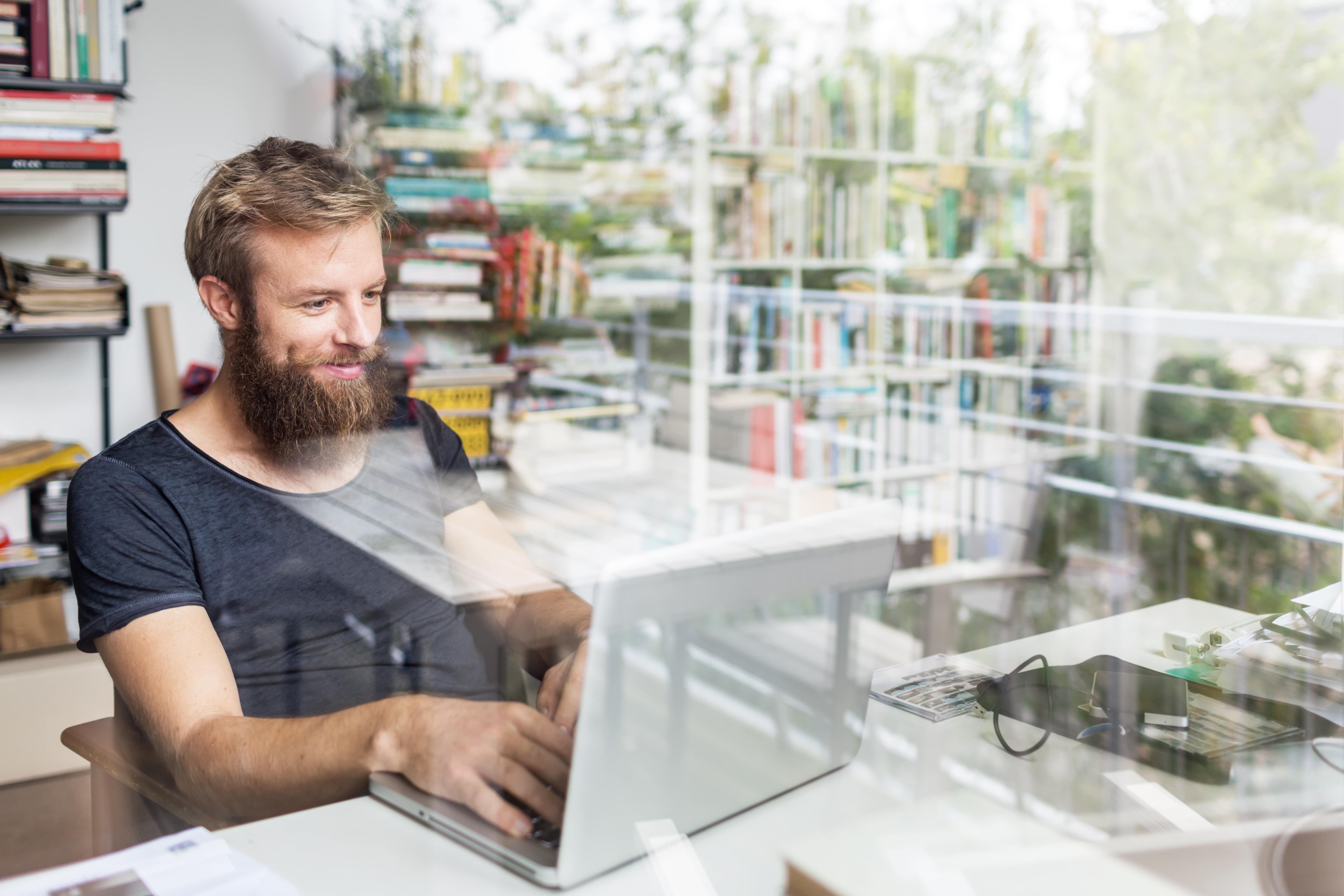 Aufnahme eines Home-Office-Schreibtisches, auf dem Notizbücher, Stifte und anderes Arbeitsmaterial liegen. Ein Mann sitzt davor, um ihn herum spielen Kinder.