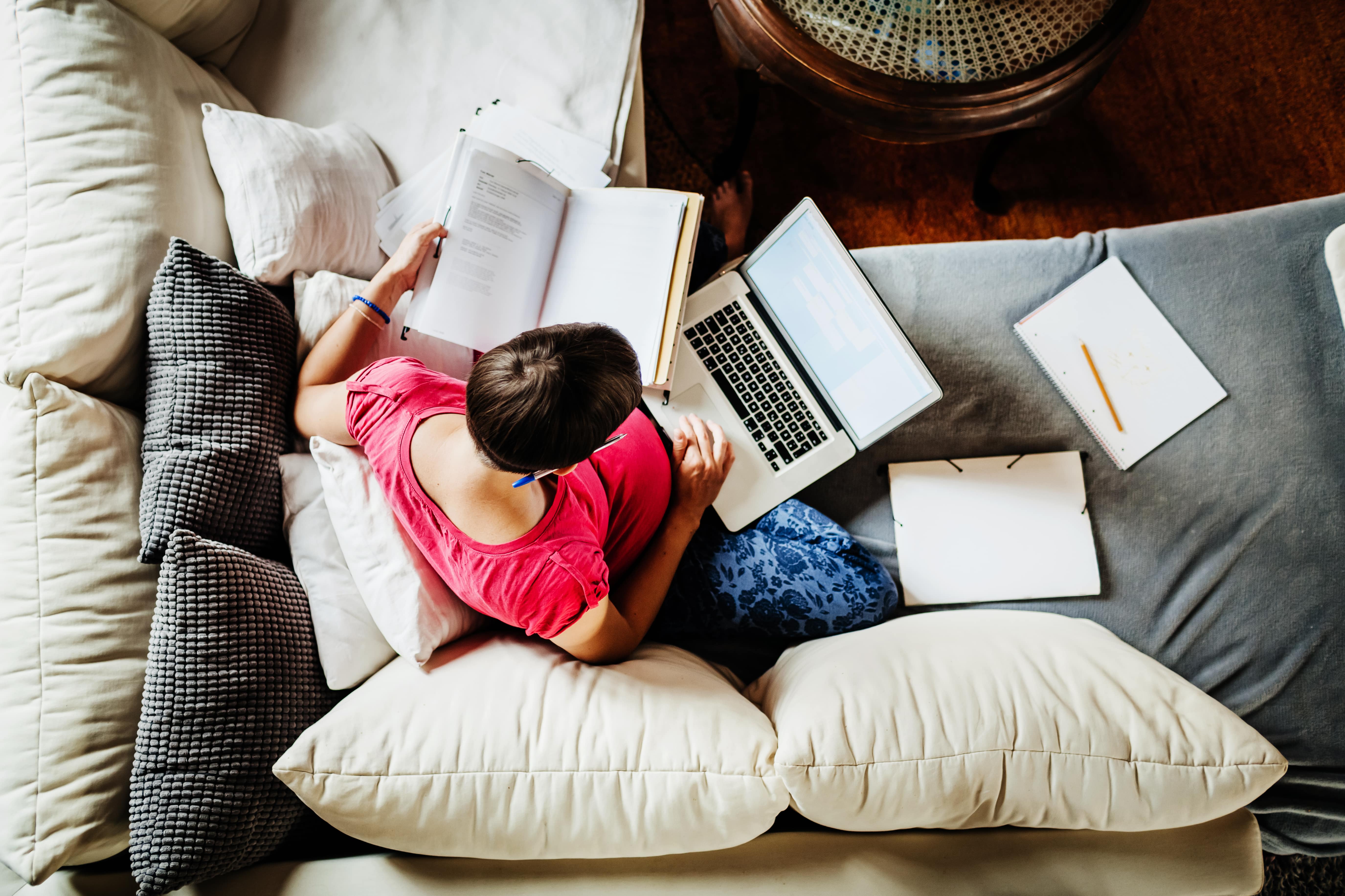 Im Home-Office sitzt eine Frau auf ihrem Sofa. Auf ihrem Schoß liegt ein Laptop, um sie herum verteilt Notizen und Hefte. Zuhause und Büro vermischen sich.
                                                                                      BU: Dein Zuhause ist nun auch dein Office? Wir zeigen dir, wie das beides geht.