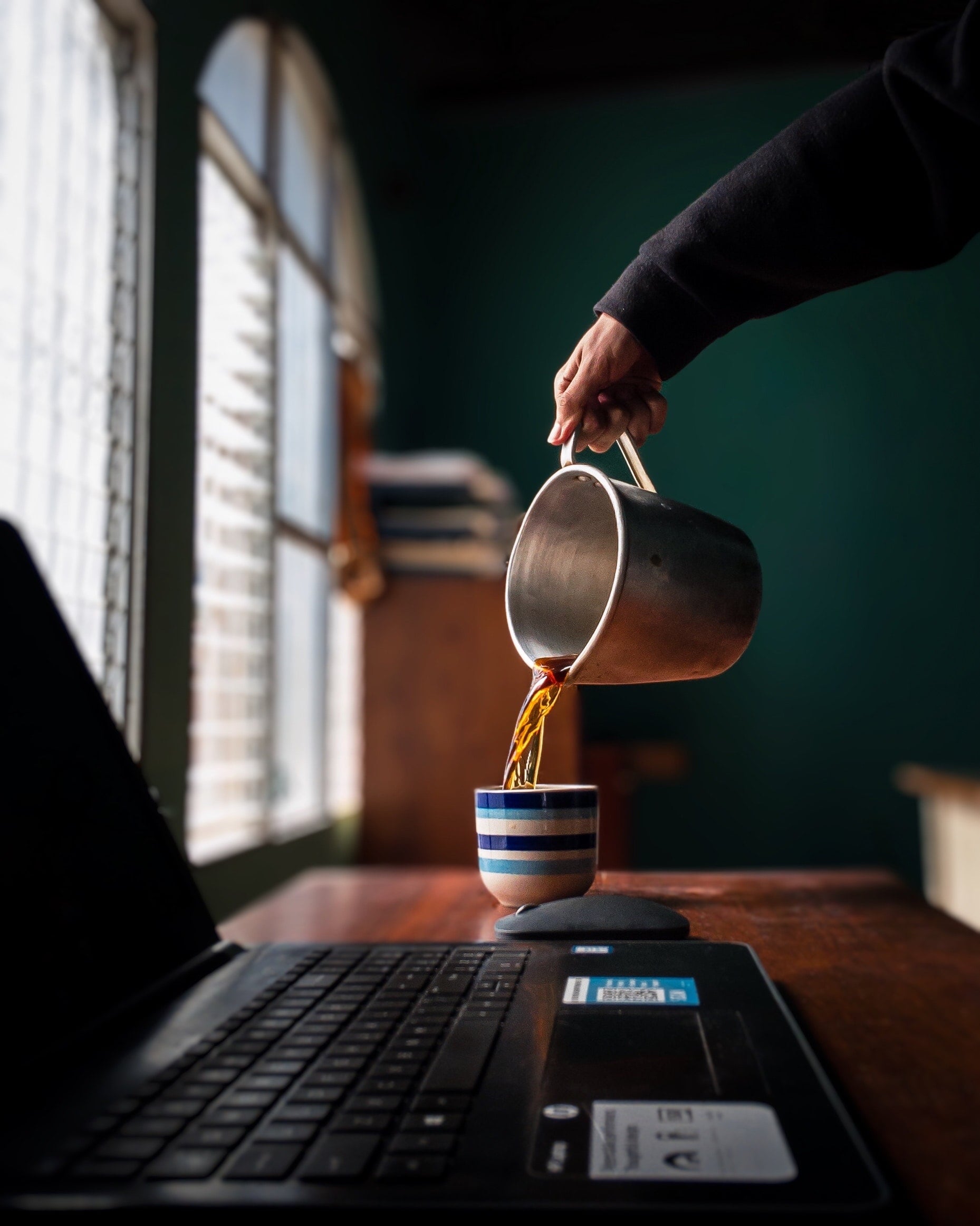 Ein Mann gießt Kaffee in eine Tasse die neben seinem Laptop auf dem Schreibtisch stehtEin Mann gießt Kaffee in eine Tasse die neben seinem Laptop auf dem Schreibtisch steht